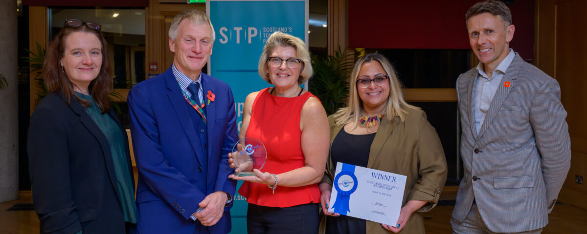 Five people pose indoors at an awards event. The person in the center holds a glass trophy, and the person next to them holds a certificate. They stand in front of a blue banner and smile at the camera.
