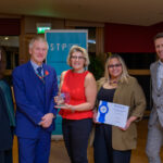 Five people pose indoors at an awards event. The person in the center holds a glass trophy, and the person next to them holds a certificate. They stand in front of a blue banner and smile at the camera.