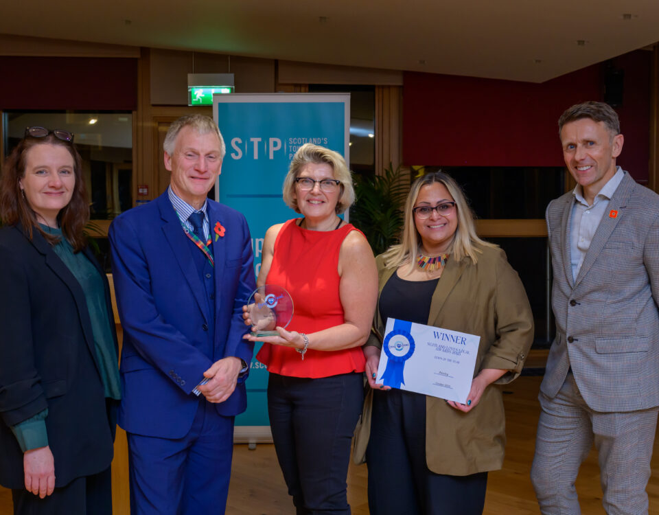 Five people pose indoors at an awards event. The person in the center holds a glass trophy, and the person next to them holds a certificate. They stand in front of a blue banner and smile at the camera.
