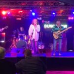 A band performs live on stage under colorful lights. Five musicians play guitars, bass, drums, and sing. Audience members watch from tables in the foreground. A sign behind the stage reads "The Bungalow.