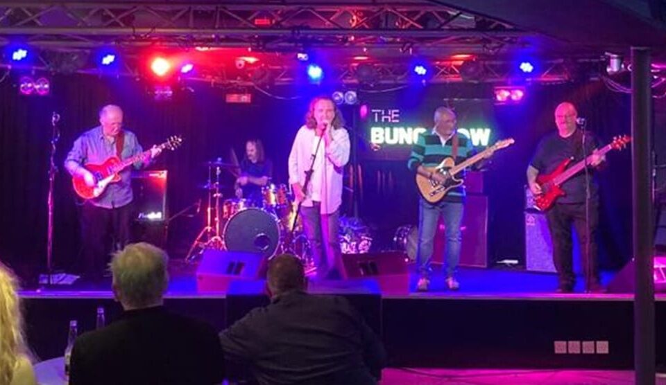 A band performs live on stage under colorful lights. Five musicians play guitars, bass, drums, and sing. Audience members watch from tables in the foreground. A sign behind the stage reads "The Bungalow.