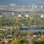 A panoramic view of a city with clusters of white buildings, residential houses, a lake, and lush greenery. High-rise buildings and hills are visible in the background under a clear sky.