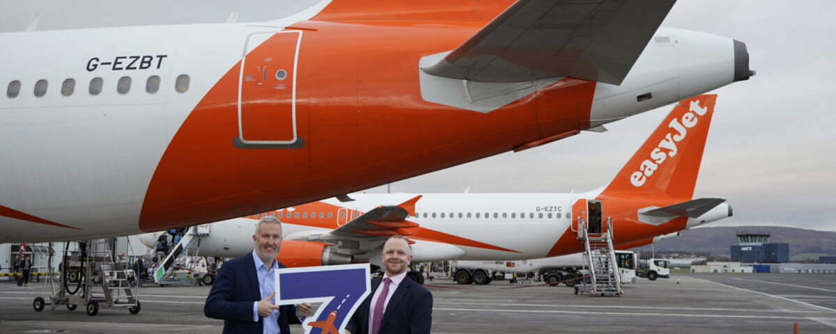 Two men in suits stand on an airport tarmac holding a large "7 easyJet" sign in front of easyJet airplanes, with the aircraft tails and engines visible in the background.