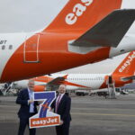 Two men in suits stand on an airport tarmac holding a large "7 easyJet" sign in front of easyJet airplanes, with the aircraft tails and engines visible in the background.