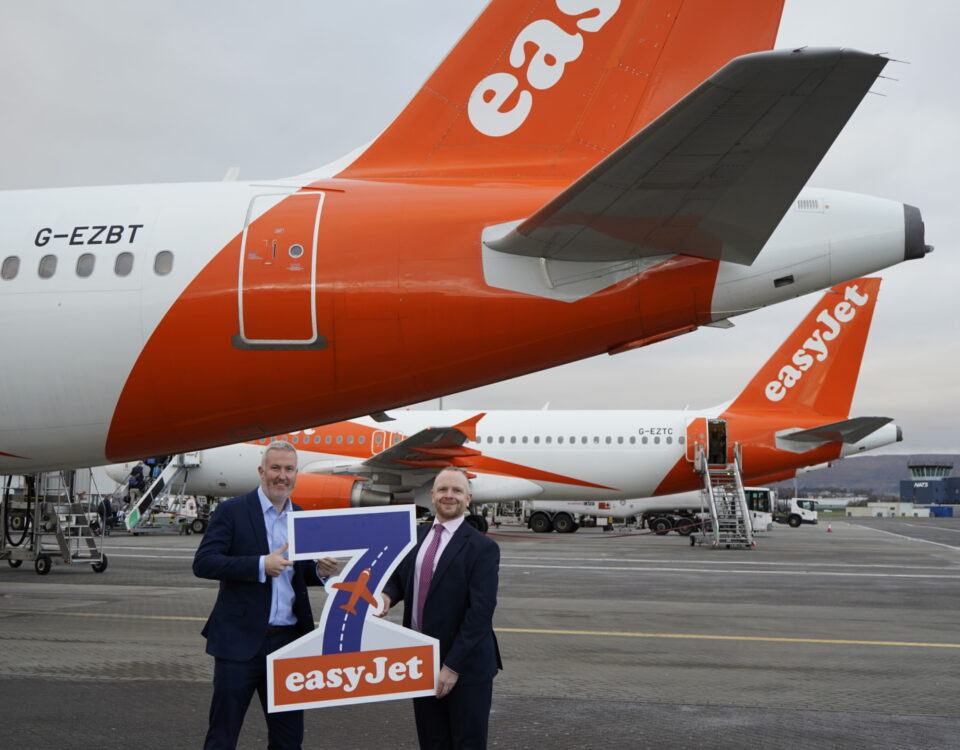 Two men in suits stand on an airport tarmac holding a large "7 easyJet" sign in front of easyJet airplanes, with the aircraft tails and engines visible in the background.