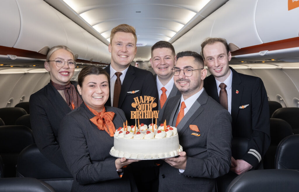 Six airline crew members in uniform stand smiling inside an airplane cabin. One holds a decorated cake with candles and a topper that says "Happy 30th Birthday." The group appears cheerful and celebratory.