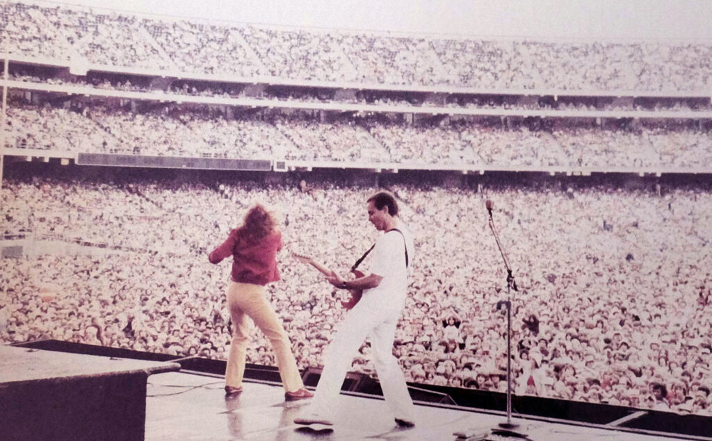 Two musicians perform energetically on a large stage in front of a massive, packed stadium crowd. One plays guitar, while the other sings or dances. The audience fills the seats and field under a bright sky.