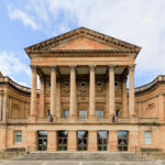 A large neoclassical building with tall columns, a triangular pediment, arched windows, and symmetrical design, set against a partly cloudy sky. Steps lead up to the entrance.