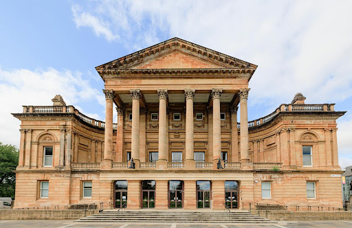A large neoclassical building with tall columns, a triangular pediment, arched windows, and symmetrical design, set against a partly cloudy sky. Steps lead up to the entrance.