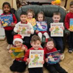 A group of young children sit and stand indoors, smiling and holding up colorful books. They are in a classroom with a wooden floor and furniture visible in the background.