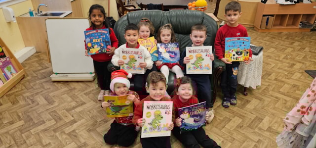 A group of young children sit and stand indoors, smiling and holding up colorful books. They are in a classroom with a wooden floor and furniture visible in the background.