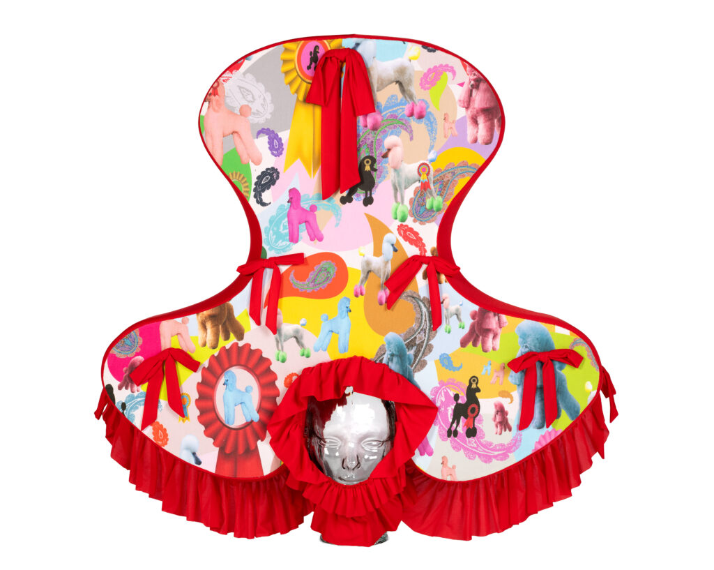 A colorful, sculptural garment with a face mask, red trim and bows, and a playful pattern of ponies, rosettes, and vibrant shapes on a white background. The design has three rounded panels and a central ruffle.