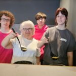 Four people stand behind a counter, three holding ladles with soup. The older woman wears glasses, and the others are younger, two in pink shirts, one in gray. Stacked bowls and a sign for cream of mushroom soup are visible.