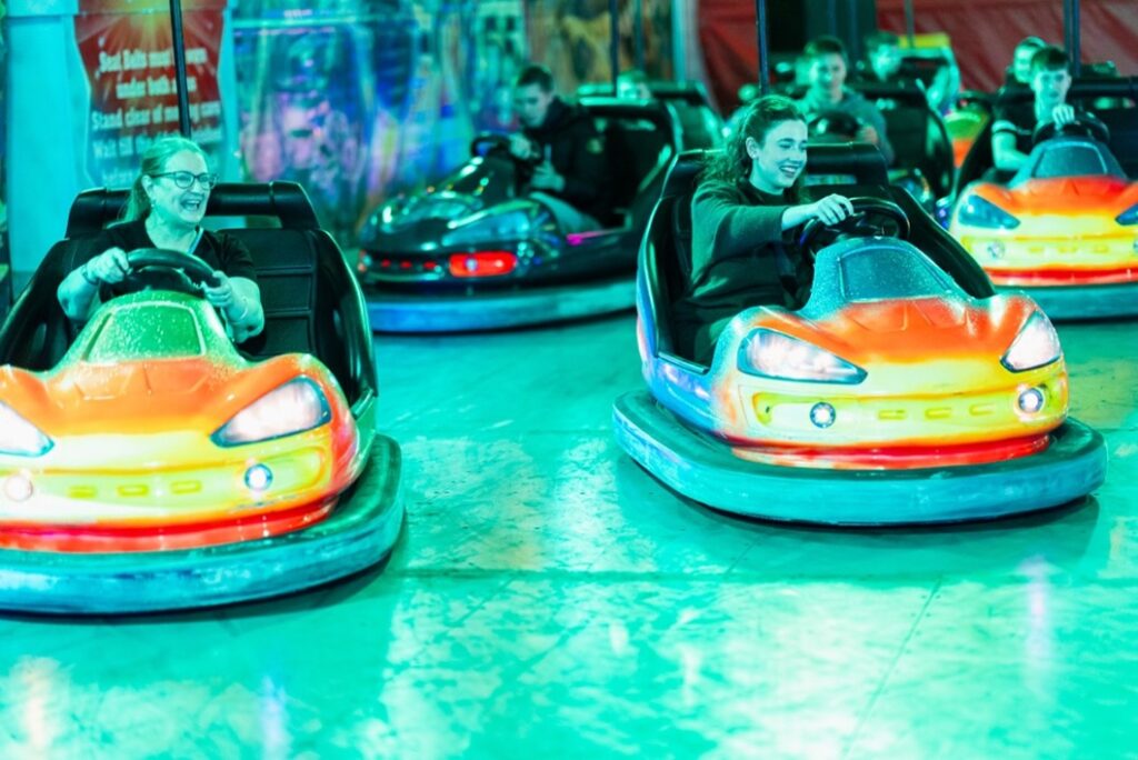 People are smiling and driving brightly colored bumper cars at an indoor amusement park, with lights creating a vibrant, playful atmosphere.
