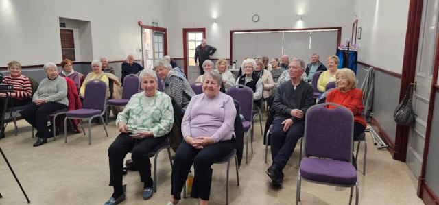 A group of older adults sit on purple chairs in a bright room, smiling and facing the camera. The room has white walls, large windows, and a few coats hanging in the background.
