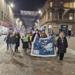 A group of people walk down a city street at night holding a blue banner and signs, participating in a march or demonstration. Buildings and streetlights line the background.