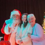 Santa Claus, an elderly woman, and a young girl in a white coat stand together, smiling beside a decorated Christmas tree with golden lights and ornaments against a dark background.