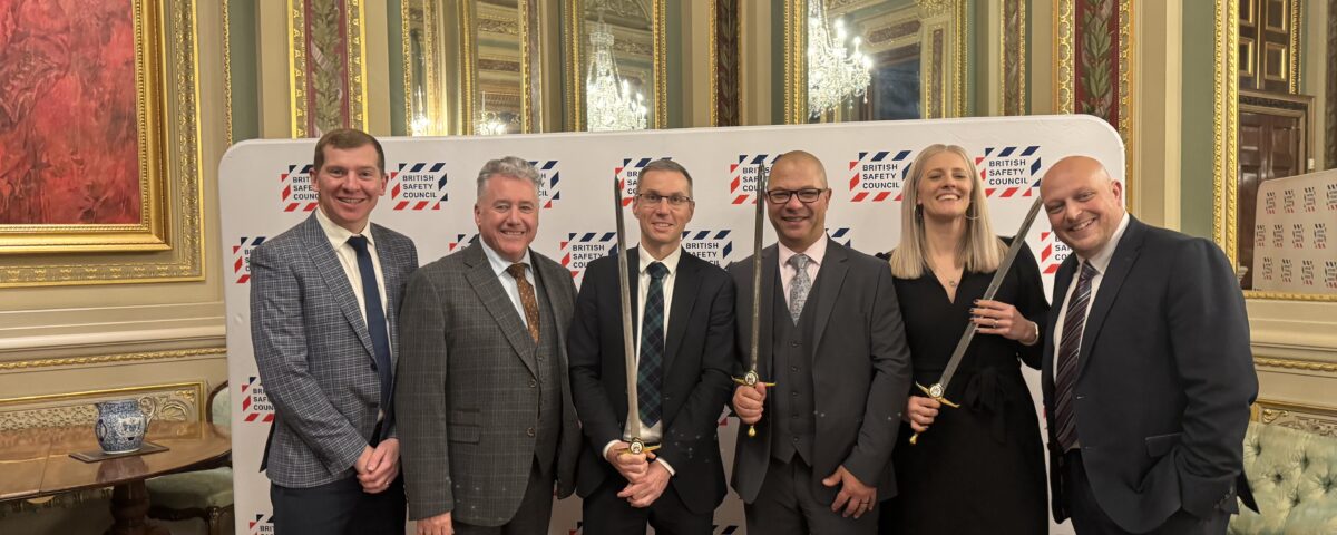 Six people in formal attire stand smiling in front of a branded backdrop in an elegant, ornate room with gold trim and mirrors. Three of them are holding ceremonial swords.