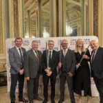 Six people in formal attire stand smiling in front of a branded backdrop in an elegant, ornate room with gold trim and mirrors. Three of them are holding ceremonial swords.
