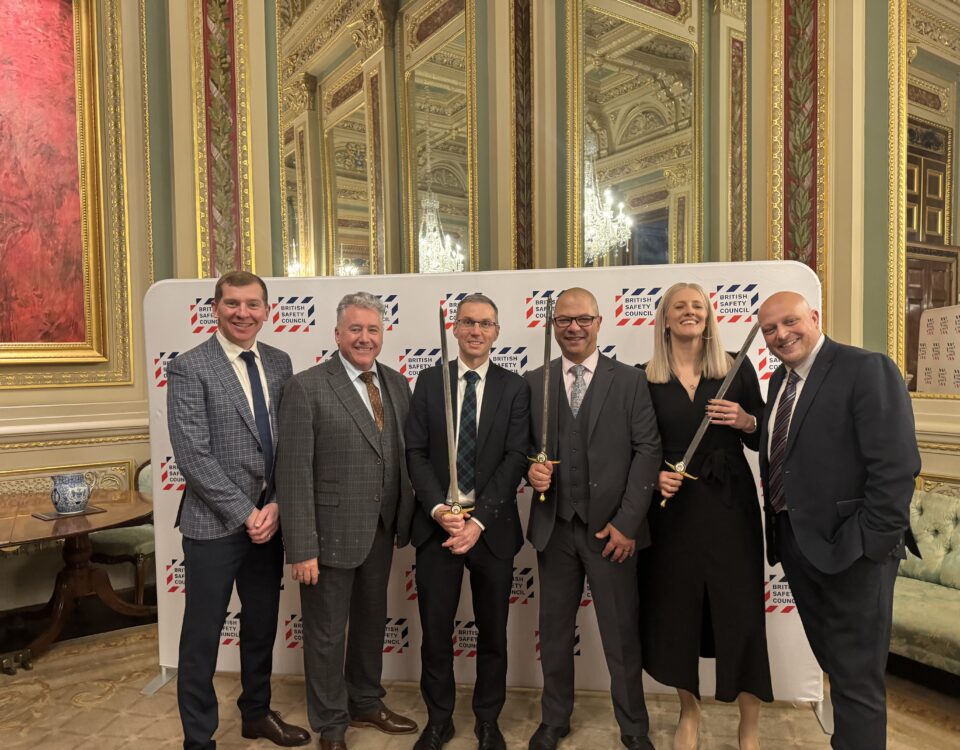 Six people in formal attire stand smiling in front of a branded backdrop in an elegant, ornate room with gold trim and mirrors. Three of them are holding ceremonial swords.