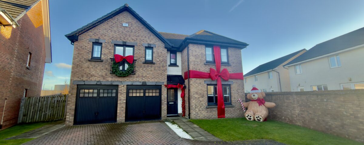 A two-story brick house with large red ribbons and a wreath, decorated for Christmas. A large inflatable teddy bear sits on the lawn next to the house, under a clear blue sky.