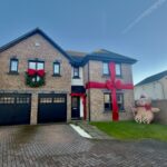 A two-story brick house with large red ribbons and a wreath, decorated for Christmas. A large inflatable teddy bear sits on the lawn next to the house, under a clear blue sky.
