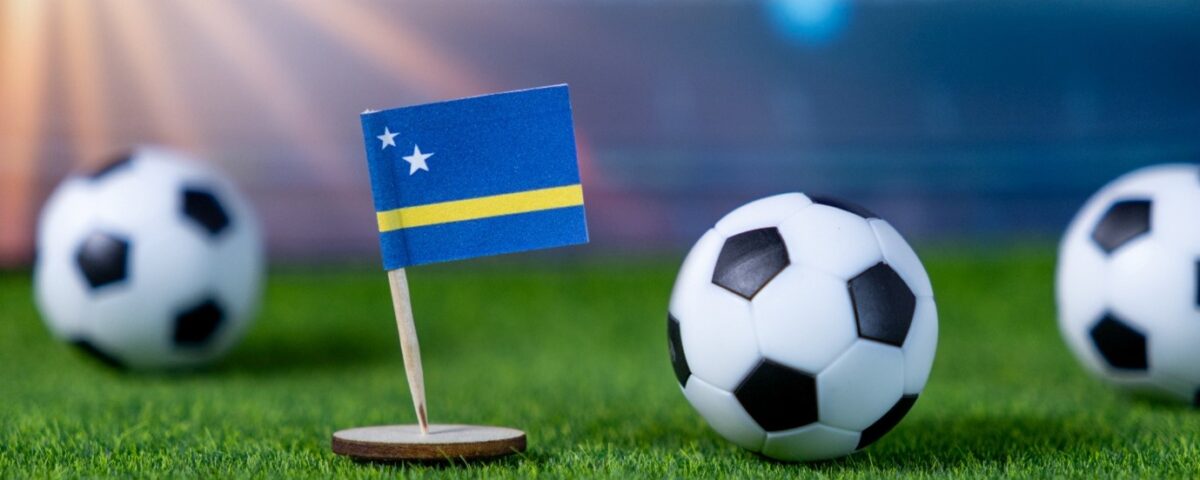 A small flag of Curaçao stands on a grassy soccer field next to a soccer ball, with two more balls blurred in the background and a stadium visible in the distance.