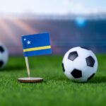 A small flag of Curaçao stands on a grassy soccer field next to a soccer ball, with two more balls blurred in the background and a stadium visible in the distance.