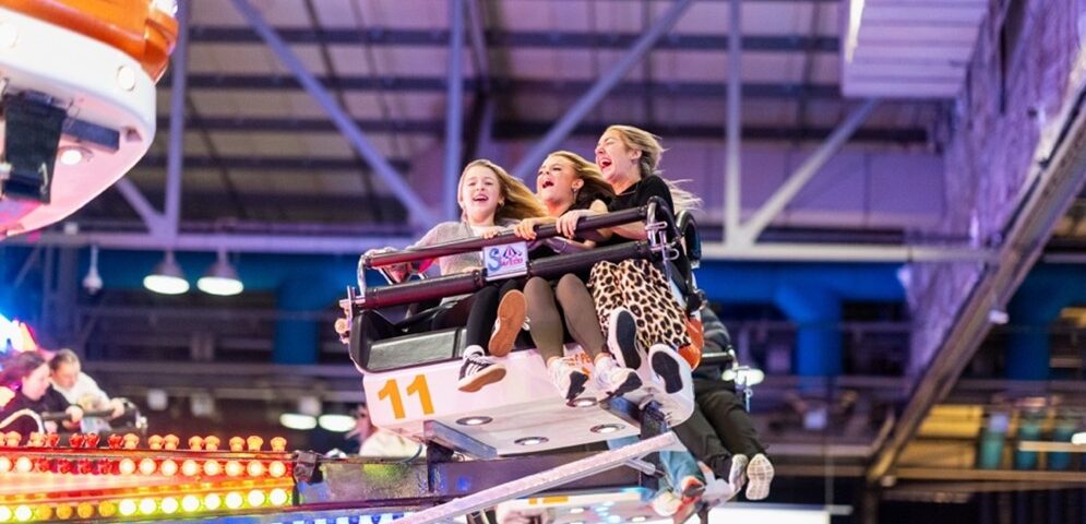 Three young women laugh and scream while riding a brightly lit amusement park ride indoors, their hair and legs flying outward as the ride spins. Colorful lights and a metal ceiling are visible in the background.