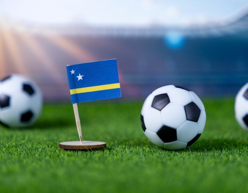 A small flag of Curaçao stands on a grassy soccer field next to a soccer ball, with two more balls blurred in the background and a stadium visible in the distance.