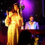 A woman in a vintage floral dress sings on stage beside a man playing piano, both under warm lights and a disco ball. A sign nearby reads "Sing a Song: A Tribute to the Carpenters.