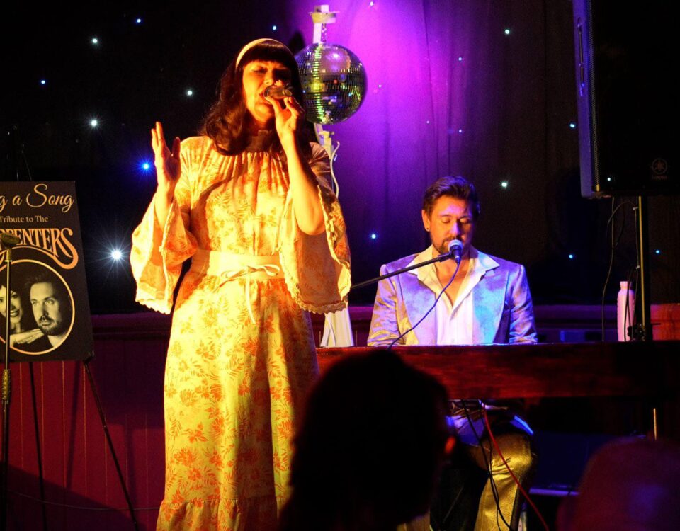 A woman in a vintage floral dress sings on stage beside a man playing piano, both under warm lights and a disco ball. A sign nearby reads "Sing a Song: A Tribute to the Carpenters.
