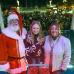 Santa Claus, a smiling girl, and a woman in a white coat pose together on a stage at a festive outdoor event with a large crowd and holiday lights in the background.