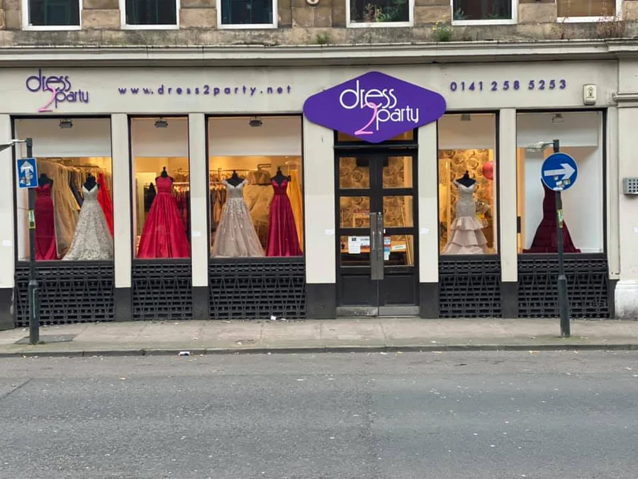 The storefront of "dress2party" displays elegant evening gowns in the window, including red, pink, and neutral-colored dresses on mannequins. The shop has large windows, a purple sign, and street signs outside.