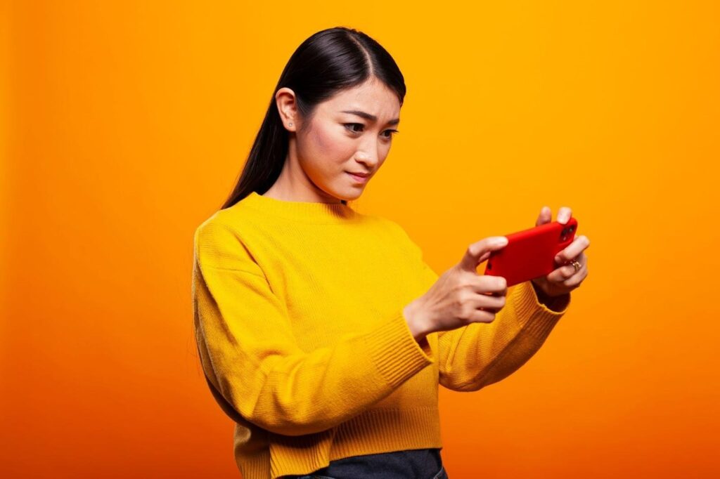 A woman in a yellow sweater holds a red smartphone with both hands, looking at the screen with a focused and slightly concerned expression, standing against an orange background.