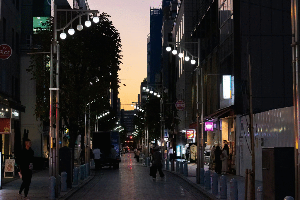 A city street at dusk is lit by rows of modern street lamps. People walk along the sidewalks, passing shops with glowing signs. The sky is a gradient of orange and blue as the sun sets behind tall buildings.