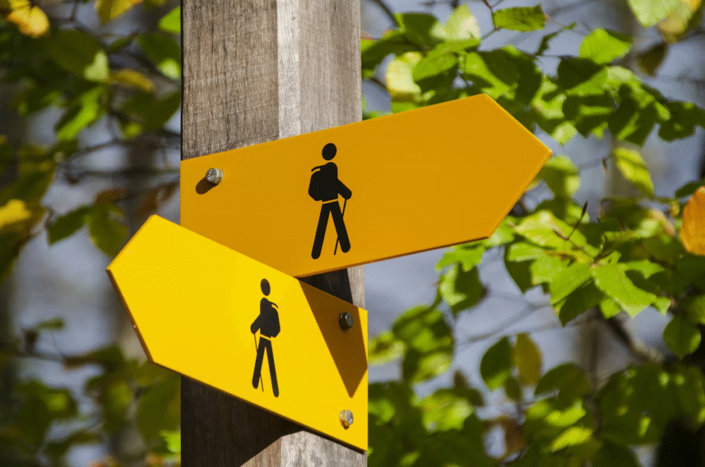 Two yellow directional signs with black hiking symbols attached to a wooden post, pointing in opposite directions. Green leaves and sunlight create a natural background.