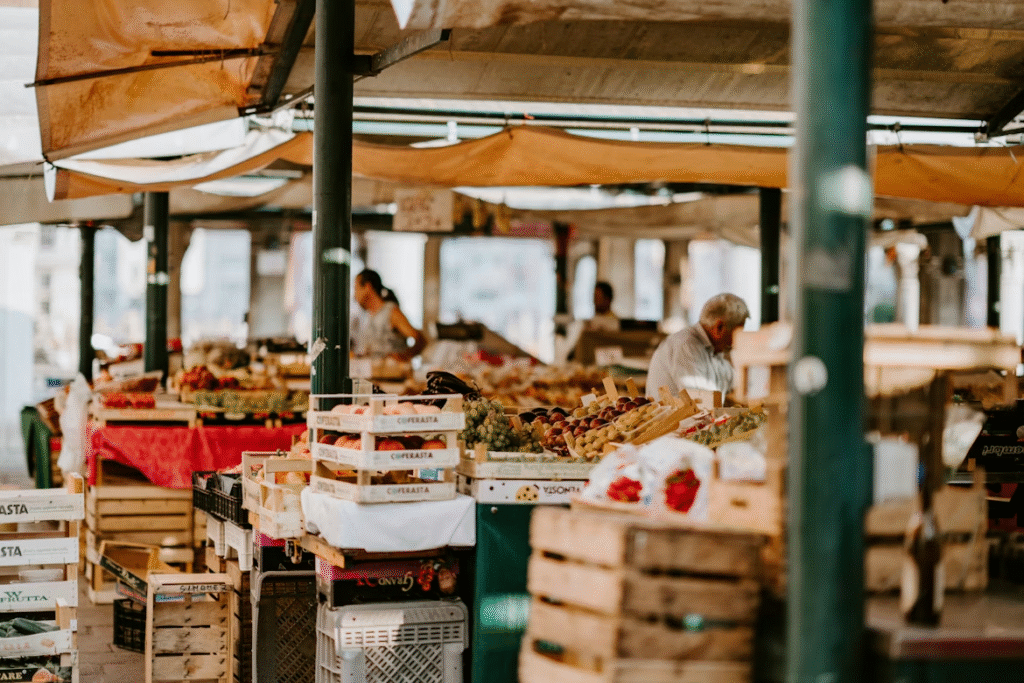 A bustling outdoor market scene with wooden crates piled high with fresh produce. Vendors and shoppers are visible in the background under a canopy, surrounded by fruits and vegetables on display.