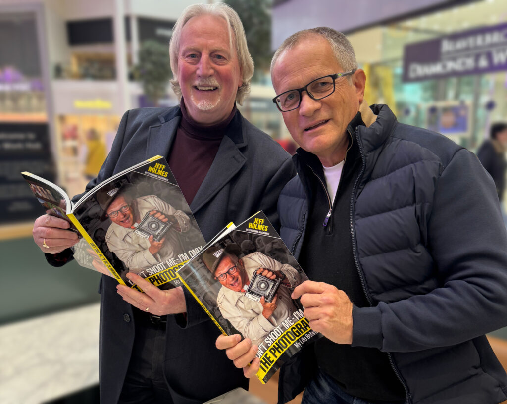 Two smiling men stand indoors holding and showing the same photography book, each with a cover photo of a man holding a camera. The background features blurred mall stores and shop signs.