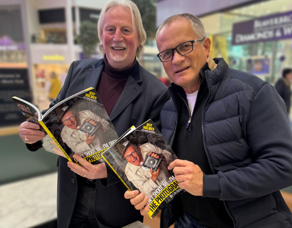 Two smiling men stand indoors holding and showing the same photography book, each with a cover photo of a man holding a camera. The background features blurred mall stores and shop signs.