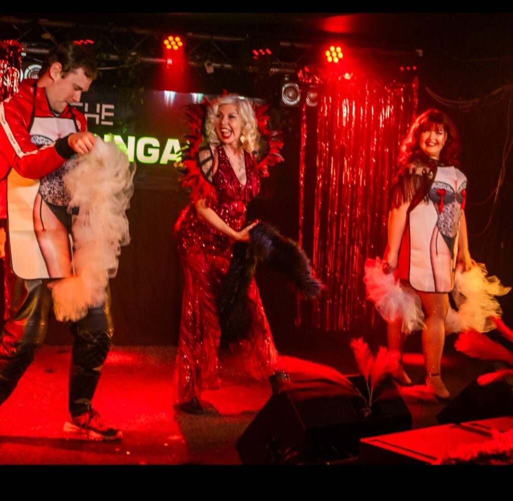 Three performers on a red-lit stage wear colorful costumes and hold feather fans. The central figure, in a sparkling red dress, smiles widely while the two flanking performers wear matching outfits with graphic prints.