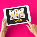 A person holds a tablet displaying a slot machine game with symbols like stars, sevens, and dollar signs on a bright pink background. The word "GAME" is written below the slot reels.