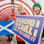 Four people stand in a colorful tunnel. Two hold a Scottish flag, while the other two hold a sign reading “CLEACHD NO CAILL: USE IT OR LOSE IT.” All are smiling and dressed for cool weather.