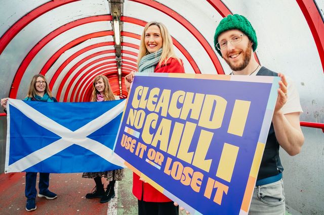 Four people stand in a colorful tunnel. Two hold a Scottish flag, while the other two hold a sign reading “CLEACHD NO CAILL: USE IT OR LOSE IT.” All are smiling and dressed for cool weather.