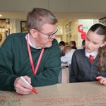 A teacher and a student sit at a table covered with notes in a classroom. The teacher points to something on the paper while the student listens attentively, both holding red markers. Other students work in the background.