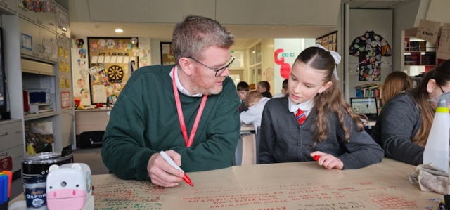 A teacher and a student sit at a table covered with notes in a classroom. The teacher points to something on the paper while the student listens attentively, both holding red markers. Other students work in the background.