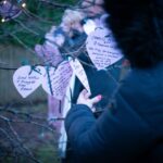 A person in a dark coat hangs a paper heart with handwritten messages on a tree. More paper hearts with notes are visible, and another person stands in the background. The scene is outdoors, with soft lighting and tree branches.