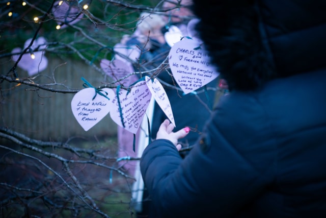 A person in a dark coat hangs a paper heart with handwritten messages on a tree. More paper hearts with notes are visible, and another person stands in the background. The scene is outdoors, with soft lighting and tree branches.