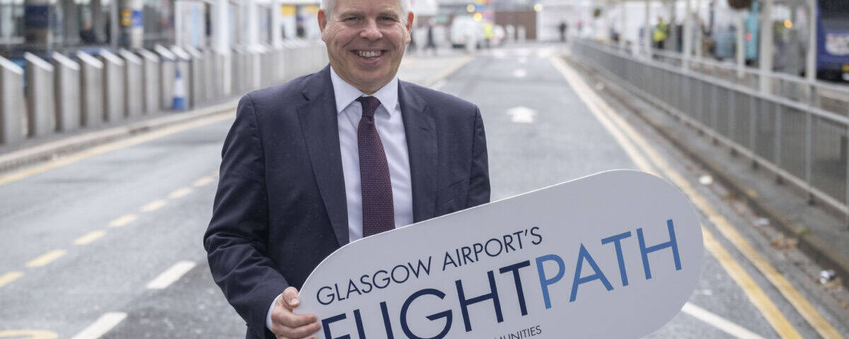 A man in a suit stands on a road outside an airport, smiling and holding a large sign that reads "Glasgow Airport's FLIGHTPATH – Proud to support our communities.