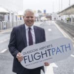 A man in a suit stands on a road outside an airport, smiling and holding a large sign that reads "Glasgow Airport's FLIGHTPATH – Proud to support our communities.